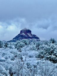Winter scene: snow-dusted red rock butte rising above frosted desert trees and shrubs under a moody cloudy sky — Southwestern U.S. landscape.