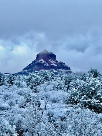 Winter scene: snow-dusted red rock butte rising above frosted desert trees and shrubs under a moody cloudy sky — Southwestern U.S. landscape.
