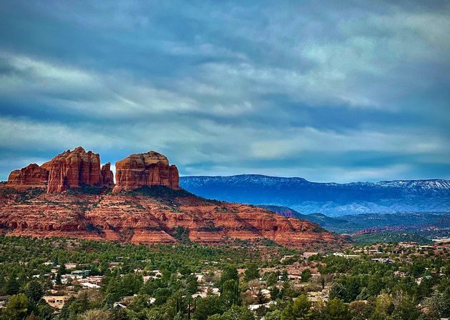 Sedona, Arizona red rock buttes towering over a green valley with scattered homes and distant snow‑dusted mountains under a moody blue sky