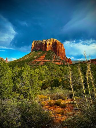 Sunlit red-rock butte rising from green desert scrub in the American Southwest, striated cliffs and pinyon-juniper forest under a dramatic deep-blue stormy sky.