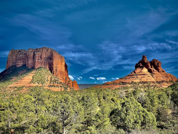 Sunlit red-rock buttes and layered sandstone formations rising above green pinyon‑juniper forest under a deep blue sky — Sedona, Arizona red rock landscape.