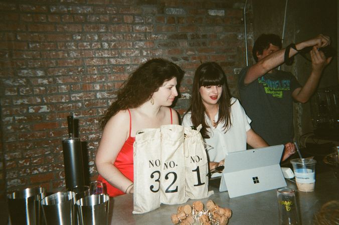 Two women at a brick-walled bar counter during a casual night out, chatting beside canvas bags labeled No. 32 and No. 1, with a tablet, metal cups, plastic drinks and a pile of champagne corks on the counter.