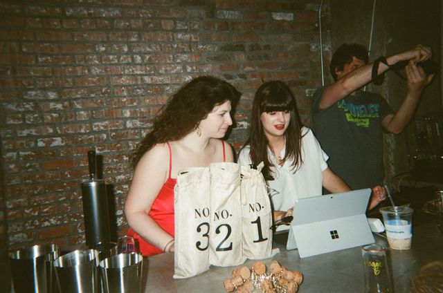 Two women at an industrial brick‑walled bar counter looking at a tablet next to canvas wine bags labeled “No. 32” and “No. 1,” metal cups and champagne corks on the bartop while someone photographs in the background.