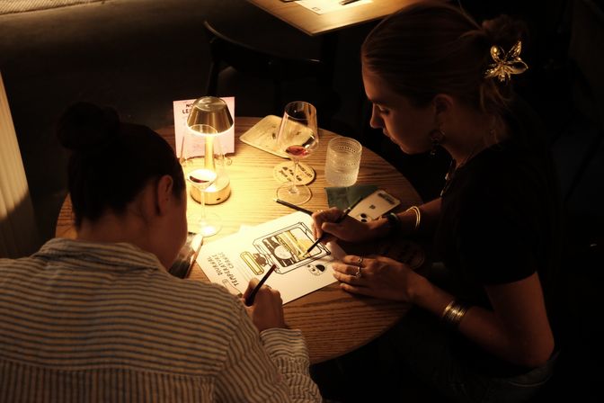 Two people leaning over sketches at a small round wooden table in a cozy dimly lit cafe, warm lamp glow, wine glasses, and drawing tools.
