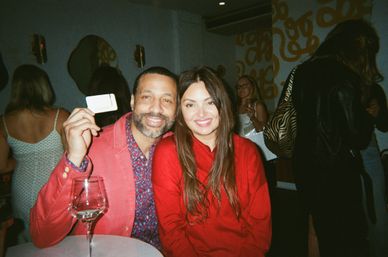 Smiling couple in a trendy urban lounge—man in coral jacket holding a card, woman in a red sweater, wine glass on the table, patterned wallpaper and other guests in the background