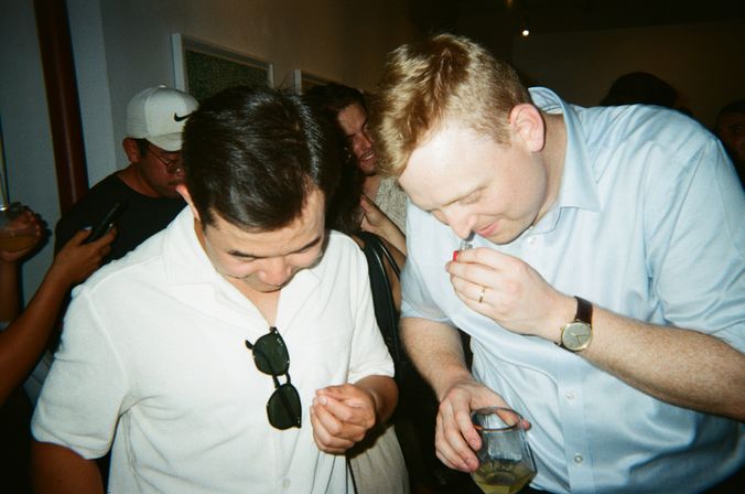Two men leaning together at a crowded indoor party tasting a sample — one sniffs a small vial while holding a cocktail, the other peers down with sunglasses clipped to his white shirt.