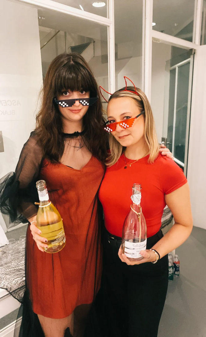 Two friends at an indoor party wearing pixelated 'deal with it' sunglasses—one with red devil horns—holding champagne bottles and dressed in red