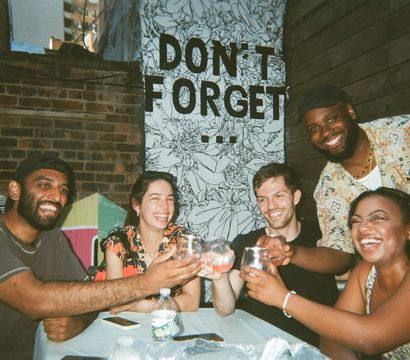 Five friends laughing and clinking glasses at an outdoor urban patio in front of a brick wall and black-and-white floral mural that reads 'DON'T FORGET'.