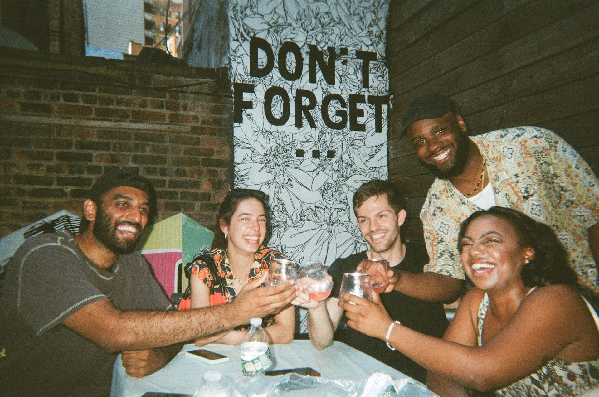 Five friends laughing and clinking glasses at an outdoor urban patio in front of a brick wall and black-and-white floral mural that reads 'DON'T FORGET'.