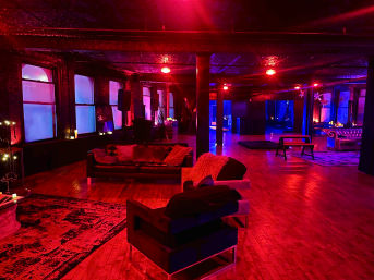 Industrial loft event space with hardwood floors, leather sofas and lounge chairs under red and blue ambient lighting, exposed columns and large windows creating a moody nightlife vibe.