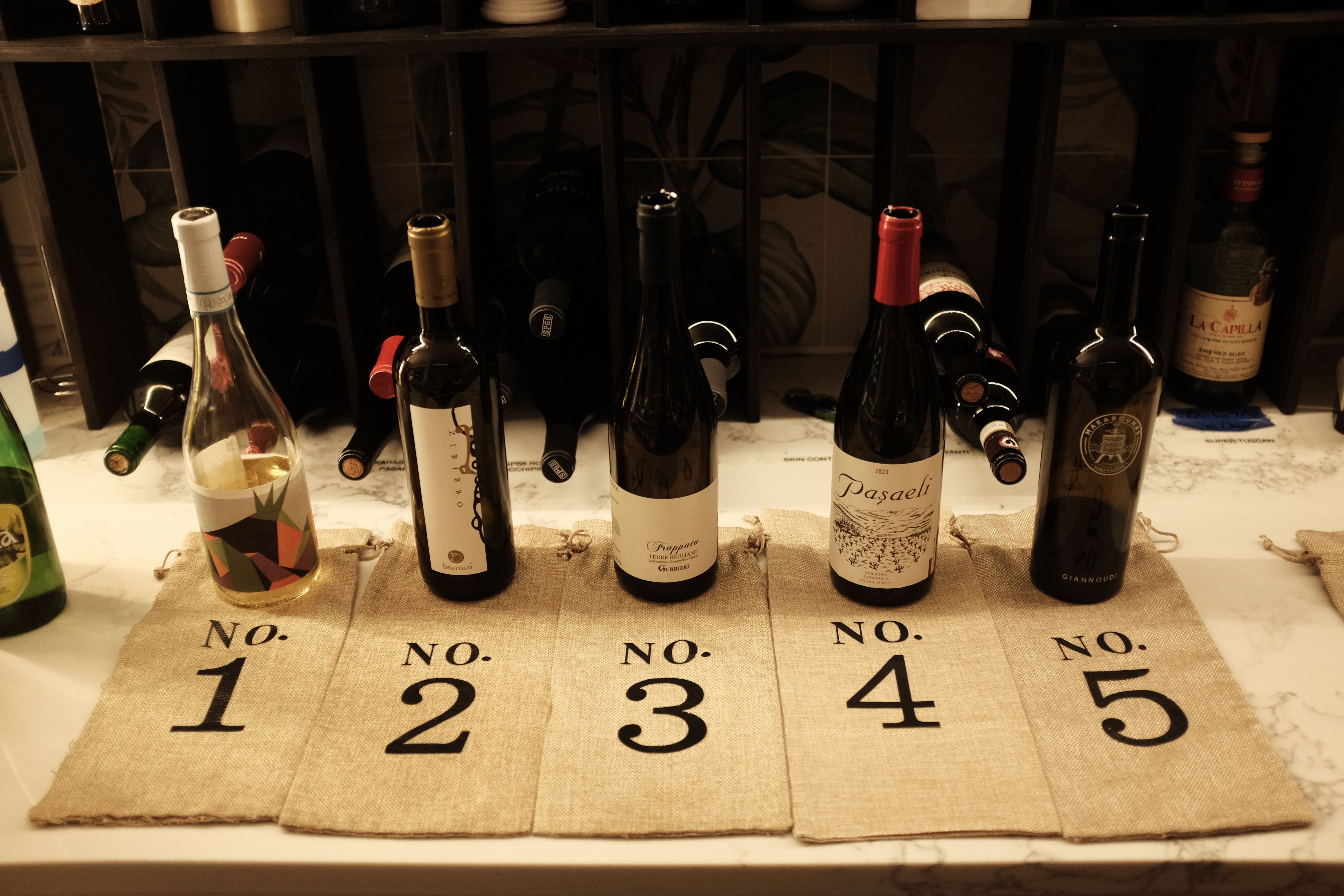 Wine tasting display on a marble countertop: five wine bottles set over numbered burlap bags No. 1–5 with a wooden wine rack behind