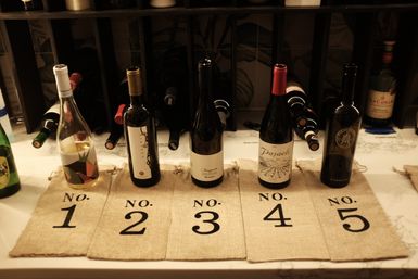 Five wine bottles lined up on a marble countertop over numbered burlap bags labeled No.1 to No.5, set in front of a wooden wine rack for a wine-tasting display.