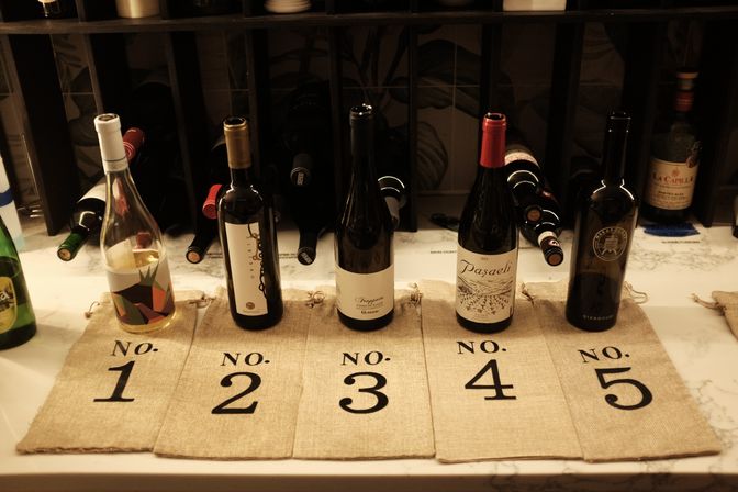 Five wine bottles lined up on a marble countertop over numbered burlap bags labeled No.1 to No.5, set in front of a wooden wine rack for a wine-tasting display.