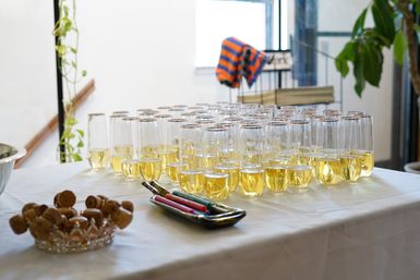 Rows of champagne flutes filled with golden sparkling wine on a white tablecloth, arranged for an indoor reception with a dish of corks and a tray of colorful markers nearby.