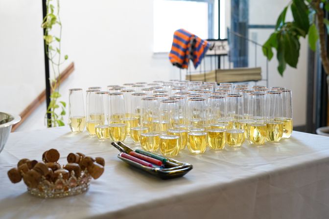 Rows of champagne flutes filled with golden sparkling wine on a white tablecloth, arranged for an indoor reception with a dish of corks and a tray of colorful markers nearby.