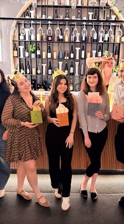 Eight friends posing at a trendy urban wine bar counter, smiling and holding colorful gift bags and wearing gold paper crowns beneath hanging wine glasses and a backlit bottle shelf.