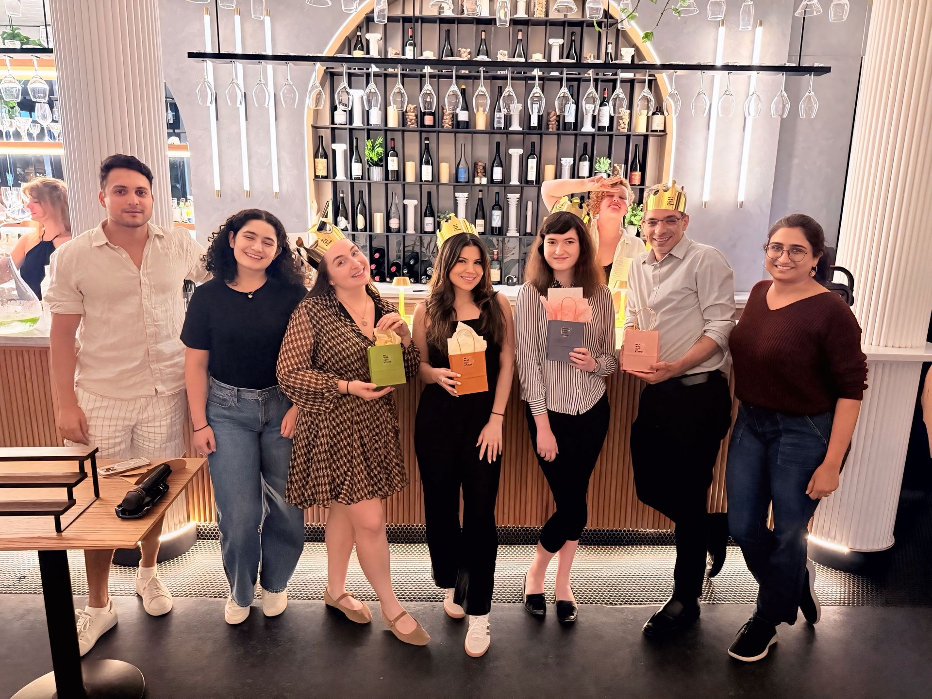 Eight friends posing at a trendy urban wine bar counter, smiling and holding colorful gift bags and wearing gold paper crowns beneath hanging wine glasses and a backlit bottle shelf.