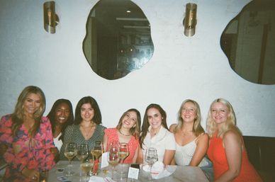 Seven friends smiling at a restaurant table enjoying wine, colorful outfits against a white brick wall with irregular mirrors and wall sconces — casual group dinner.