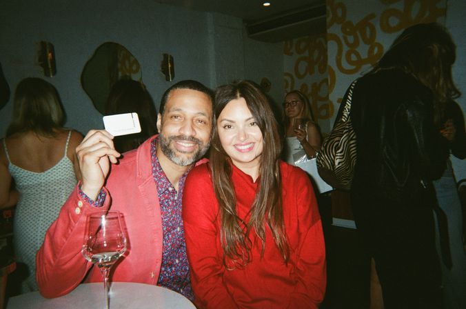 Smiling man and woman in red outfits seated at a lively indoor cocktail party — man holds a small card above a wine glass on a round table, other guests and patterned wall art in the background.