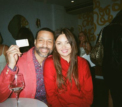 Smiling friends in red outfits pose at a lively indoor lounge — man holds a card, wine glass on the table, guests and a yellow-patterned wall in the background.