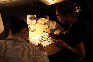 Two people at a small round wooden table in a cozy dimly lit cafe, coloring a printed illustration with markers while wine glasses, a water tumbler, a phone, and a warm tabletop lamp sit nearby.