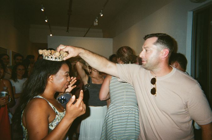 Man places a decorative crown on a smiling woman’s head at a lively indoor art gallery party, guests mingling and watching in the background.