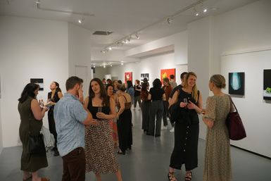 Guests mingling at a contemporary art gallery opening, people in casual attire holding drinks and chatting in a white‑walled exhibition space with paintings on display.