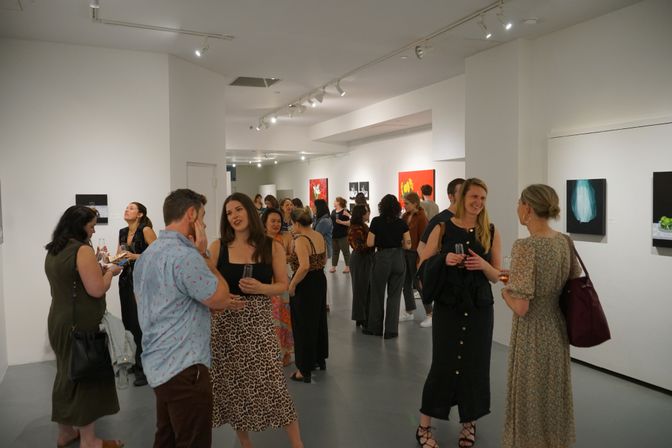 Guests mingling at a contemporary art gallery opening, people in casual attire holding drinks and chatting in a white‑walled exhibition space with paintings on display.