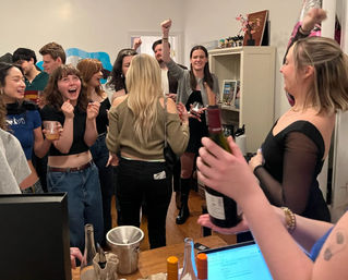Lively apartment house party in a cozy living room, friends cheering and laughing while raising wine bottles and glasses.