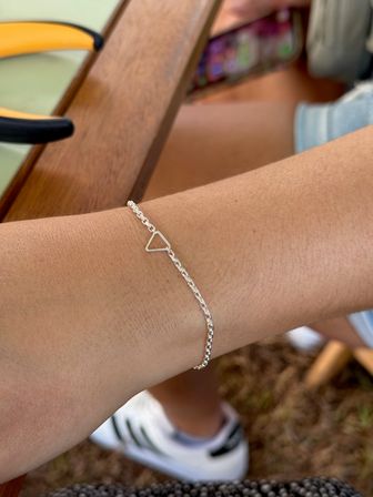 Close-up of a wrist wearing a delicate silver chain bracelet with a tiny triangle charm, resting on a wooden table at an outdoor patio with sneakers and grass in the background.