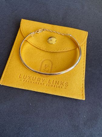 Shiny gold bangle bracelet with small safety chain resting on a mustard-yellow suede jewelry pouch, flatlay on dark textured fabric