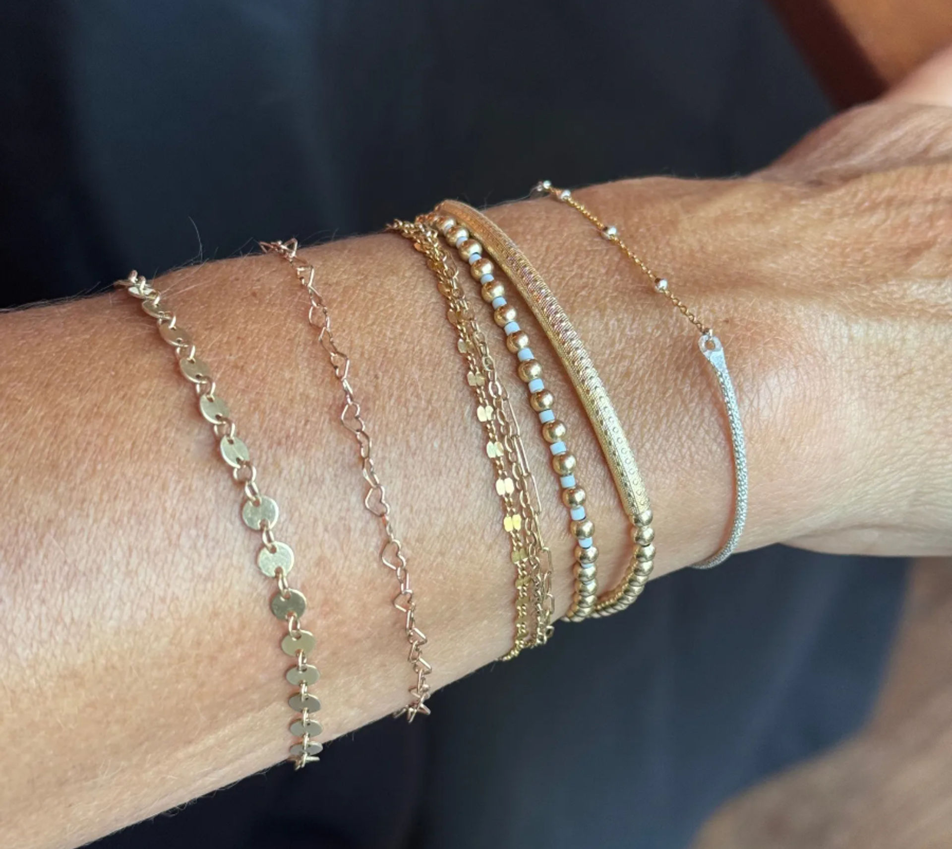 Close-up of a wrist wearing stacked dainty gold and silver bracelets with mixed chains, tiny beads, and textured bars against a dark background.
