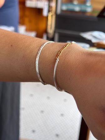 Close-up of a wrist wearing two delicate bracelets — a patterned silver bangle and a thin gold chain with a silver tube detail, blurred indoor background.