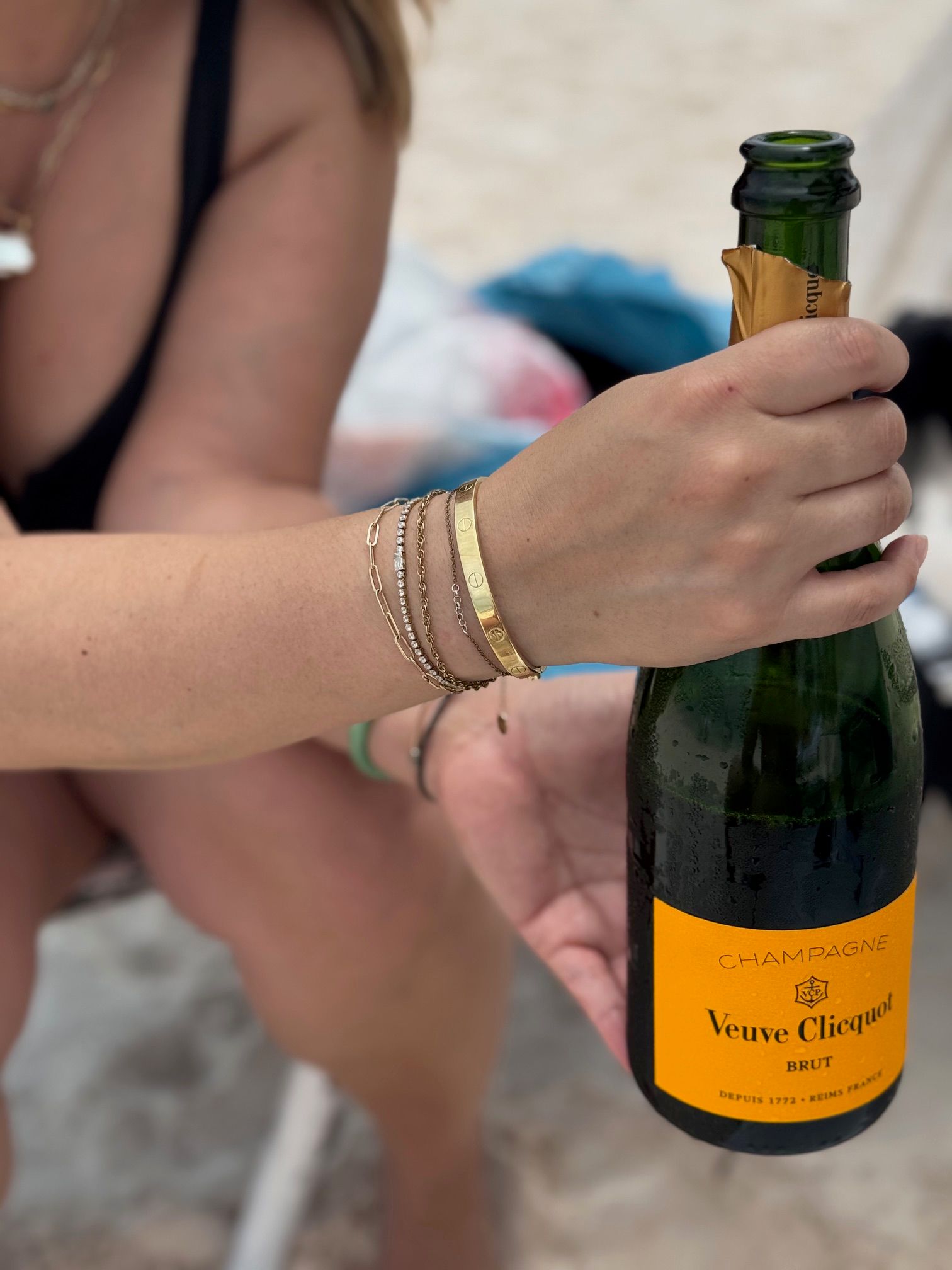 Close-up of a hand with gold bracelets opening a chilled champagne bottle with a bright yellow-orange label on a sandy beach, summer celebration vibe.