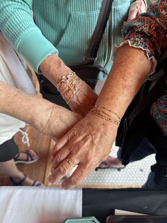 Close-up of three hands wearing stacked gold bracelets and a small green-stone ring over a jewelry display at a local craft market