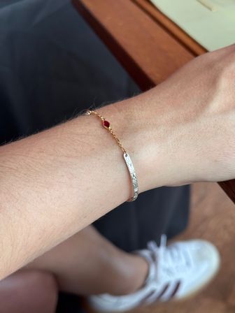 Close-up of a wrist wearing minimalist jewelry: a delicate gold chain with a tiny red gemstone and a slim engraved silver bangle, resting by a wooden table with white sneakers blurred in the background.