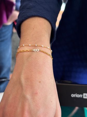 Close-up of a wrist wearing two dainty gold bracelets: a tiny pearl chain and a thin link chain with a small silver 'B' initial charm against a navy sleeve background.