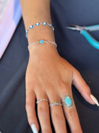 Close-up of a hand and wrist wearing a delicate silver chain bracelet with a small blue stone and a beaded evil-eye bracelet, with stacked rings including a large turquoise oval ring, resting on dark fabric next to turquoise-handled jewelry pliers.