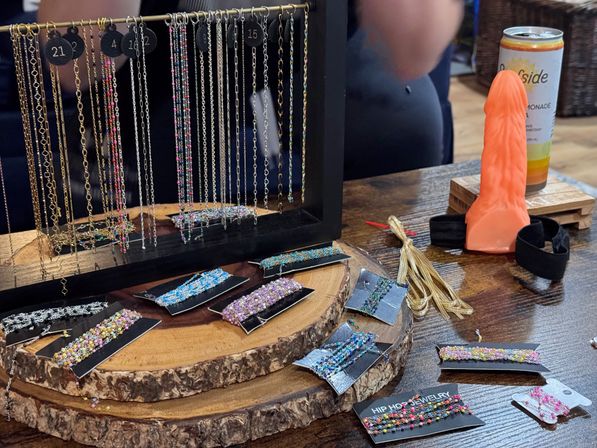 Artisan beaded necklaces and bracelets hanging from a black frame and displayed on rustic wood slabs at a craft fair table, with an orange silicone novelty adult toy and a canned drink at the side.