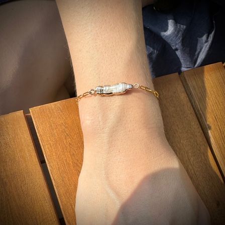 Close-up of a sunlit wrist resting on wooden slats, wearing a delicate gold chain bracelet with a wrapped irregular freshwater pearl centerpiece — outdoor patio jewelry detail.