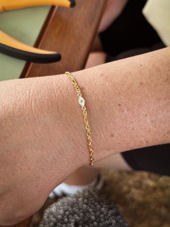Close-up of a wrist wearing a delicate gold chain bracelet with a tiny eye-shaped charm and gemstone, resting on a wooden patio table — casual summer accessory shot