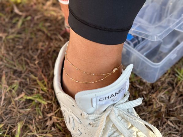 Close-up of an ankle wearing two delicate gold anklets with a crescent charm tucked into a white sneaker on dry grass in an outdoor casual setting