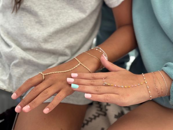 Two hands side by side with pastel pink and mint nails, wearing delicate gold chain hand-harnesses and beaded bracelets on a casual fabric background.