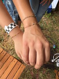 Close-up of two hands stacked outdoors showing dainty gold chain bracelets with tiny colorful beads and a black-and-white checkered paper wristband, grass and wooden slats in the background.