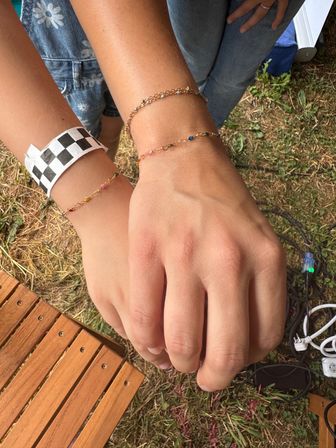 Close-up of two hands stacked outdoors showing dainty gold chain bracelets with tiny colorful beads and a black-and-white checkered paper wristband, grass and wooden slats in the background.