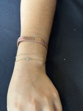 Close-up of a wrist wearing a textured copper cuff and two delicate gold chain bracelets, one with a small blue gemstone, resting on a dark fabric background.