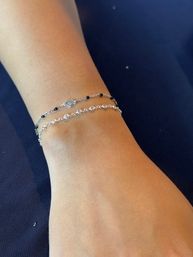 Close-up of a wrist wearing two delicate silver bracelets — a thin chain with tiny black beads and heart charm and a sparkling linked crystal chain — resting on navy fabric.