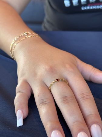 Close-up of a manicured hand with French-tip nails resting on navy fabric, wearing a delicate gold chain ring with a small red gemstone on the middle finger and stacked gold bracelets.