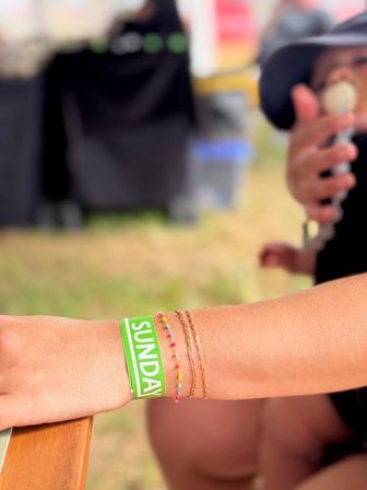 Sunlit wrist wearing a green "SUNDAY" event wristband with colorful beaded and gold bracelets, blurred background of a person eating ice cream at an outdoor summer festival