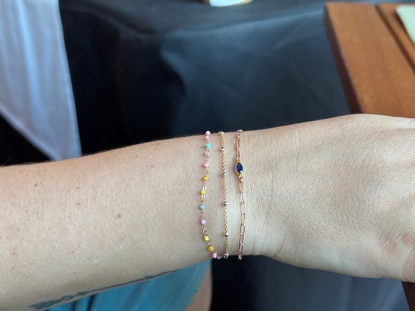 Close-up of a wrist wearing three delicate stacked bracelets: playful pastel beaded strand, thin gold beaded chain, and rose-gold paperclip chain with a small blue marquise gemstone on a neutral background